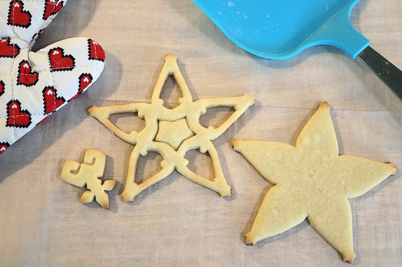 Pu&ograve; includere: Vista dall'alto di biscotti a forma di stella di varie dimensioni, accanto a un guanto da forno rosso e bianco con un motivo a cuore e una spatola blu. I biscotti sono dorati e il guanto ha uno sfondo bianco con cuori pixelati rossi.