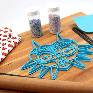 May include: A blue plastic cookie cutter shaped like the Majora's Mask from the Legend of Zelda video game. The cutter is on a wooden cutting board with a rolling pin, a spatula, and two jars of sprinkles.