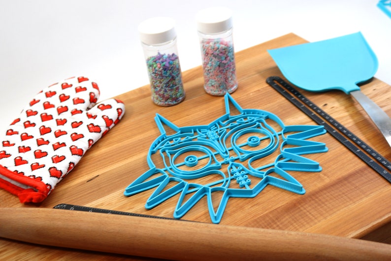 May include: A blue plastic cookie cutter shaped like the Majora's Mask from the Legend of Zelda video game. The cutter is on a wooden cutting board with a rolling pin, a spatula, and two jars of sprinkles.