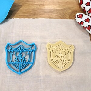 May include: Two cookie cutters in the shape of a shield with a design of a bird and a triangle inside. One cutter is blue plastic and the other is beige and appears to be made of dough.