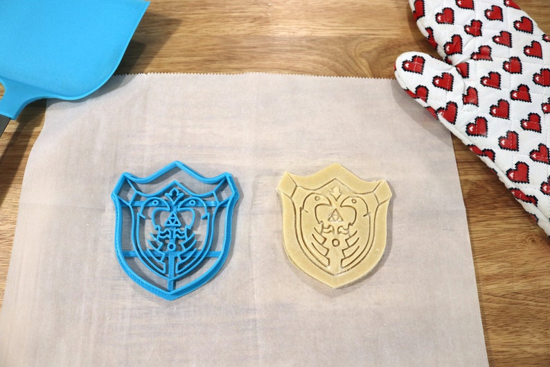 May include: Two cookie cutters in the shape of a shield with a design of a bird and a triangle inside. One cutter is blue plastic and the other is beige and appears to be made of dough.