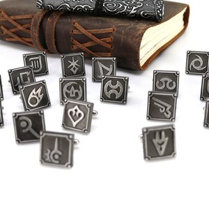 May include: A set of 18 silver cufflinks with different symbols, including a sword, a shield, a fire, and a star. The cufflinks are arranged on a white surface.