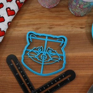 Puede incluir: Un cortador de galletas de pl&aacute;stico azul con forma de cara de mapache. El cortador tiene un dise&ntilde;o detallado con un contorno negro.