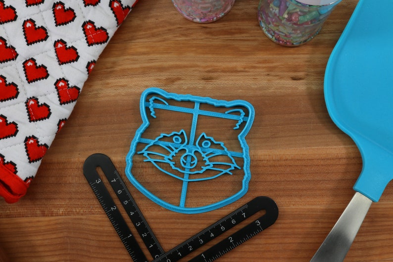 Puede incluir: Un cortador de galletas de pl&aacute;stico azul con forma de cara de mapache. El cortador tiene un dise&ntilde;o detallado con un contorno negro.