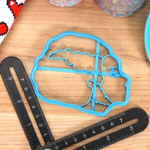 May include: A light blue plastic cookie cutter in the shape of a sailboat. A black ruler and a white cloth with red pixelated hearts are also visible. The items are on a wooden surface.