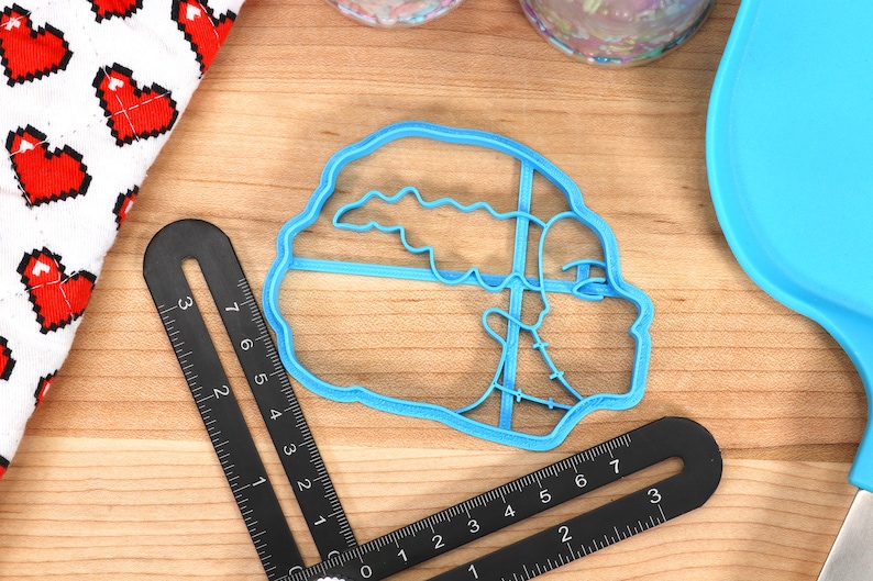 May include: A light blue plastic cookie cutter in the shape of a sailboat. A black ruler and a white cloth with red pixelated hearts are also visible. The items are on a wooden surface.