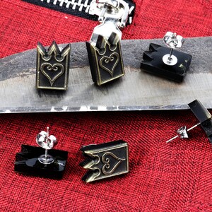 May include: Earrings with a crown and heart design. The earrings are black and gold, with a post backing. The earrings are displayed on a metal surface with a red fabric background.