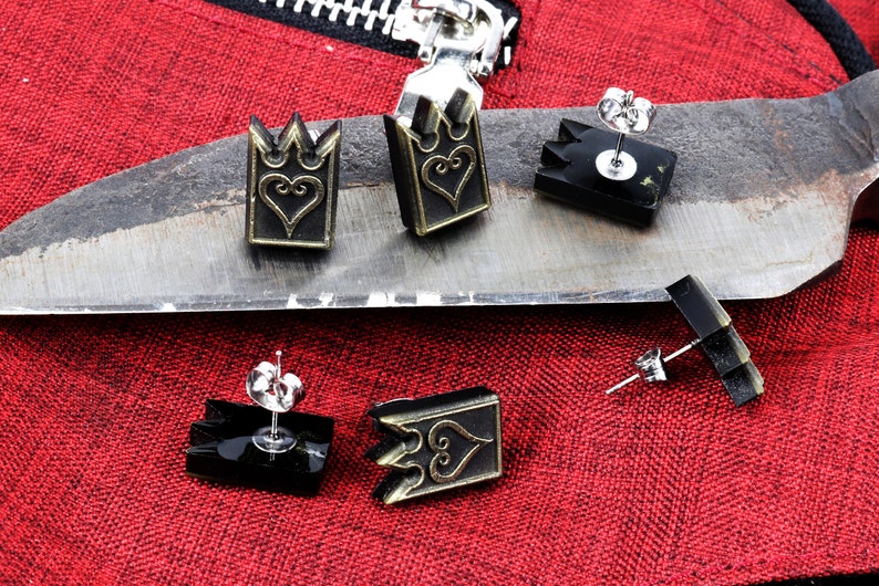 May include: Earrings with a crown and heart design. The earrings are black and gold, with a post backing. The earrings are displayed on a metal surface with a red fabric background.