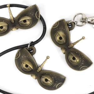 May include: Three gold-colored robot eye charms, one on a black cord necklace and two on a silver key chain. The charms have a detailed design with two large eyes and a small antenna.