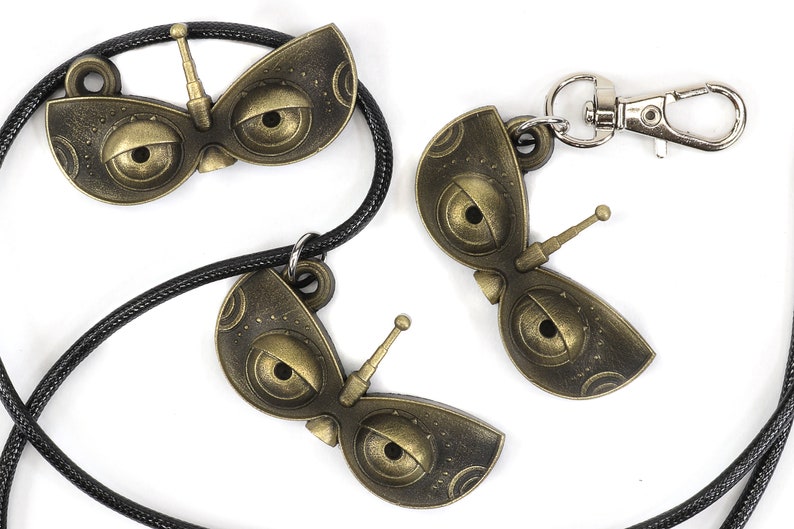 May include: Three gold-colored robot eye charms, one on a black cord necklace and two on a silver key chain. The charms have a detailed design with two large eyes and a small antenna.