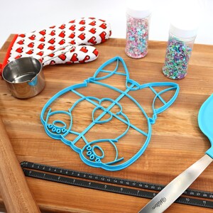 May include: A blue plastic cookie cutter shaped like a dog's face, a wooden rolling pin, a small metal measuring cup, a pair of red oven mitts with white hearts, two jars of colorful sprinkles, a blue silicone spatula, and a black ruler.