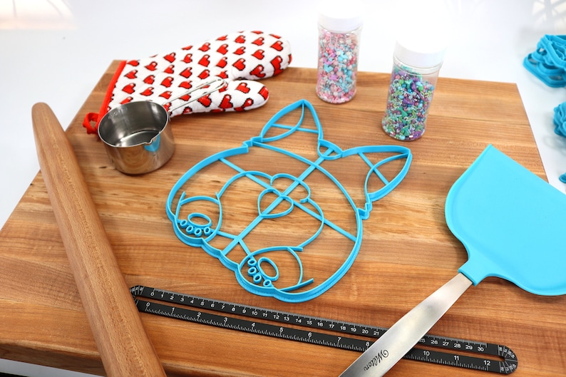 May include: A blue plastic cookie cutter shaped like a dog's face, a wooden rolling pin, a small metal measuring cup, a pair of red oven mitts with white hearts, two jars of colorful sprinkles, a blue silicone spatula, and a black ruler.