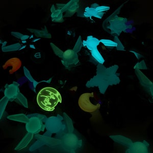 May include: A collection of glow-in-the-dark charms featuring various shapes and designs, including a crescent moon, stars, and a character with a surprised expression.