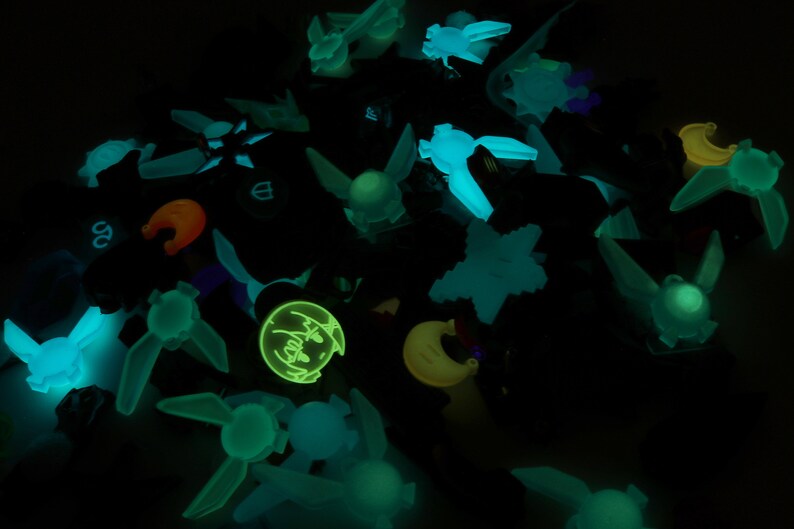 May include: A collection of glow-in-the-dark charms featuring various shapes and designs, including a crescent moon, stars, and a character with a surprised expression.