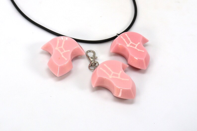 May include: Three pink resin pendants with a white Gemini zodiac symbol. The pendants are attached to a black cord necklace and a silver key chain.