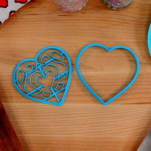 May include: Two blue heart-shaped cookie cutters on a wooden surface. One cutter is plain, while the other has a decorative design.