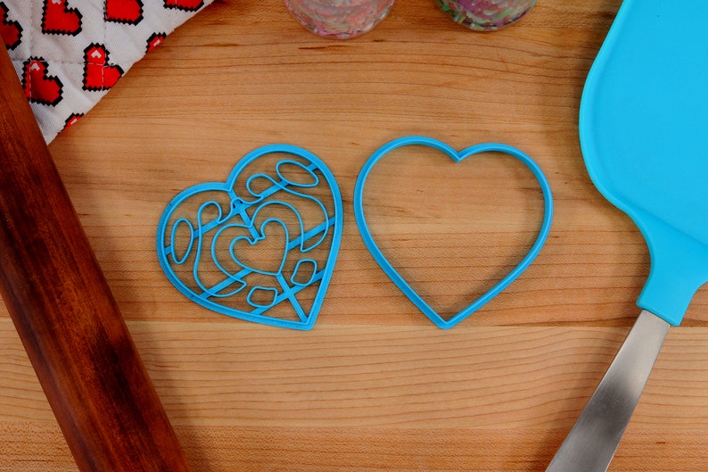 May include: Two blue heart-shaped cookie cutters on a wooden surface. One cutter is plain, while the other has a decorative design.