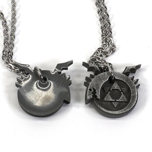 K&ouml;nnte beinhalten: Zwei silberne Halsketten mit Anh&auml;ngern, die die alchemistischen Symbole f&uuml;r die Elemente Feuer und Erde darstellen. Das Feuersymbol ist ein Dreieck mit einer nach oben zeigenden Spitze, und das Erdsymbol ist ein Dreieck mit einer nach unten zeigenden Spitze.