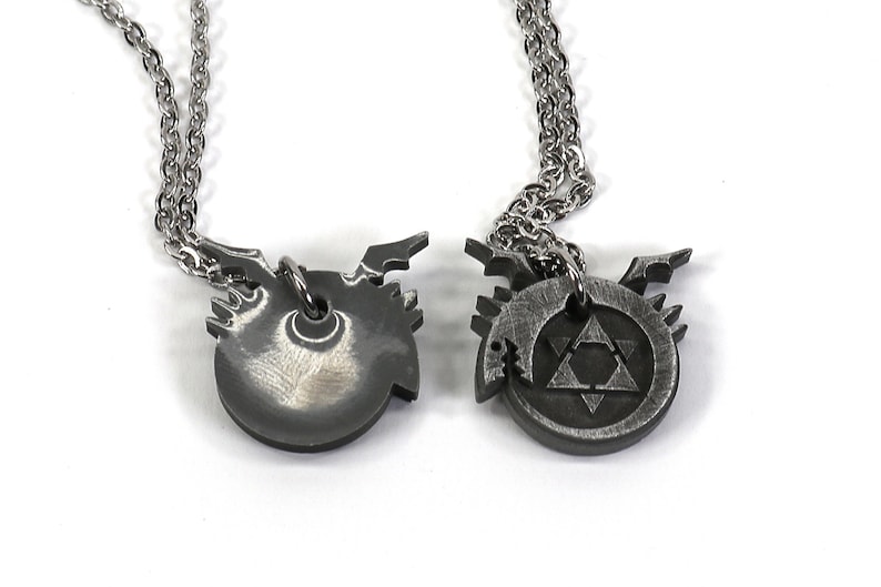 K&ouml;nnte beinhalten: Zwei silberne Halsketten mit Anh&auml;ngern, die die alchemistischen Symbole f&uuml;r die Elemente Feuer und Erde darstellen. Das Feuersymbol ist ein Dreieck mit einer nach oben zeigenden Spitze, und das Erdsymbol ist ein Dreieck mit einer nach unten zeigenden Spitze.