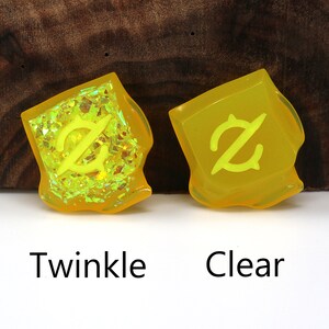 May include: Two yellow resin dice with a symbol on each face. The dice on the left has a glittery finish, while the dice on the right is clear. The dice are labeled "Twinkle" and "Clear".