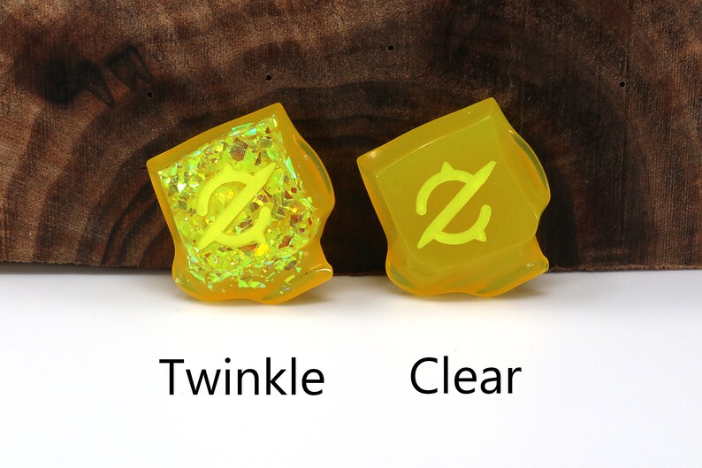 May include: Two yellow resin dice with a symbol on each face. The dice on the left has a glittery finish, while the dice on the right is clear. The dice are labeled "Twinkle" and "Clear".