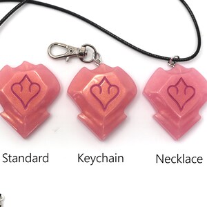 May include: Three pink resin pendants with a heart-shaped design, labeled "Standard", "Keychain", and "Necklace". The pendants are attached to a black cord and a silver keychain clasp. The background is white.