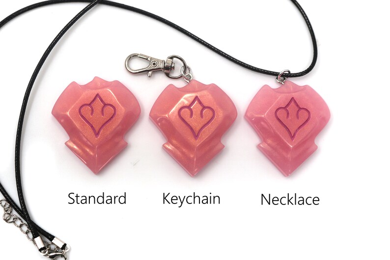 May include: Three pink resin pendants with a heart-shaped design, labeled "Standard", "Keychain", and "Necklace". The pendants are attached to a black cord and a silver keychain clasp. The background is white.