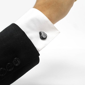 May include: A black suit jacket with buttons on the cuff. The cuff is folded back to reveal a pair of silver cufflinks shaped like a teardrop.