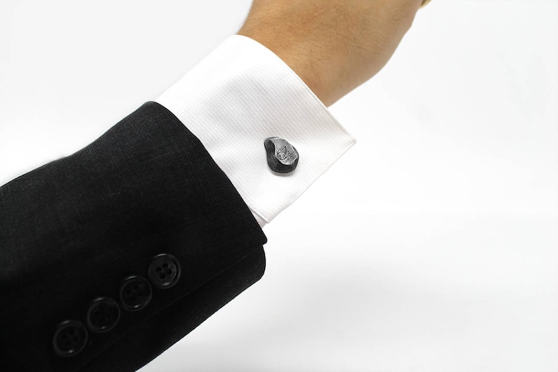 May include: A black suit jacket with buttons on the cuff. The cuff is folded back to reveal a pair of silver cufflinks shaped like a teardrop.