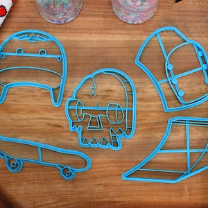 May include: A set of five blue plastic cookie cutters shaped like a skateboard, a skull, a beanie, a ramp, and a shirt. The cutters are on a wooden surface.