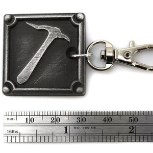 May include: A square, metal keychain with a pickaxe design. The pickaxe is silver against a dark gray background. The keychain has a silver clasp and a ring to attach keys. The square is approximately 1 inch by 1 inch.