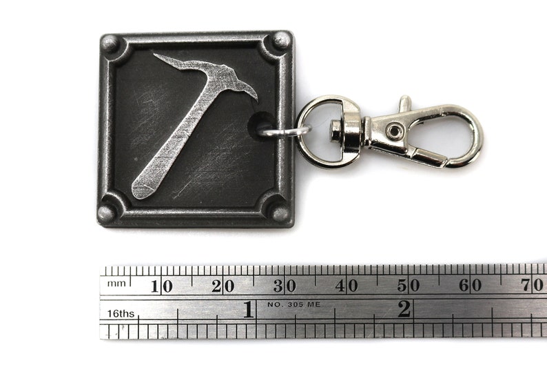 May include: A square, metal keychain with a pickaxe design. The pickaxe is silver against a dark gray background. The keychain has a silver clasp and a ring to attach keys. The square is approximately 1 inch by 1 inch.