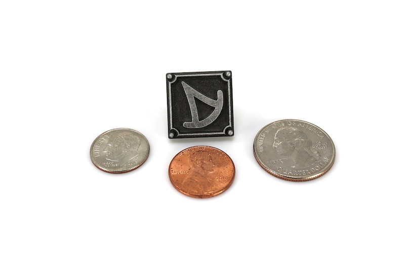 May include: A silver metal pin with a black background and a stylized silver symbol. The pin is surrounded by three US coins, a penny, a dime, and a quarter.