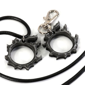Puede incluir: Dos anillos de metal de color plateado con un dise&ntilde;o dentado en forma de engranaje. Un anillo est&aacute; unido a un collar de cord&oacute;n negro, y el otro anillo est&aacute; unido a un llavero plateado.