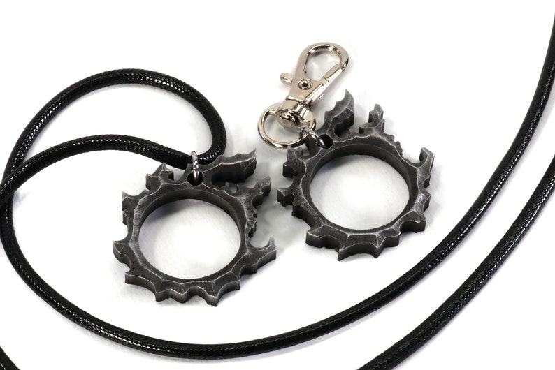 Puede incluir: Dos anillos de metal de color plateado con un dise&ntilde;o dentado en forma de engranaje. Un anillo est&aacute; unido a un collar de cord&oacute;n negro, y el otro anillo est&aacute; unido a un llavero plateado.