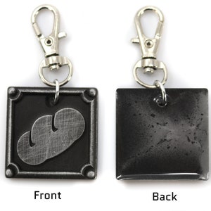 May include: A black metal keychain with a silver-colored mushroom design on the front and a plain black back. The keychain has a silver-colored metal clasp.