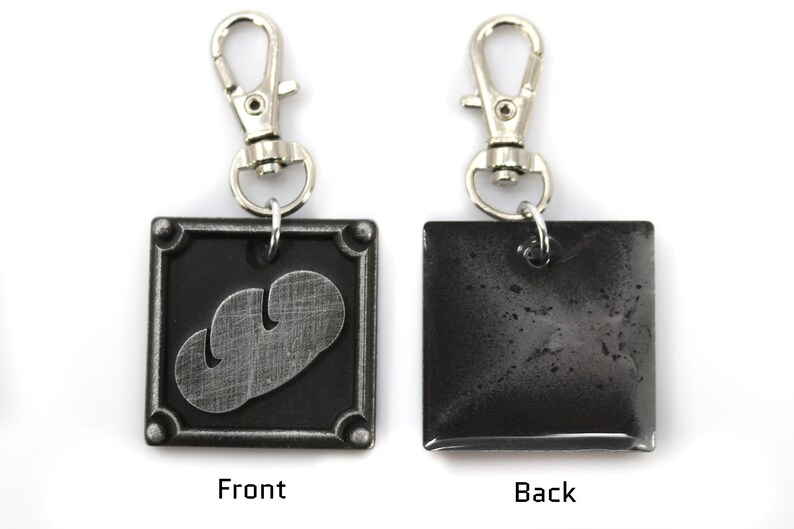 May include: A black metal keychain with a silver-colored mushroom design on the front and a plain black back. The keychain has a silver-colored metal clasp.