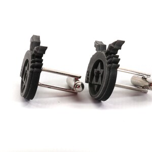 May include: A pair of black cufflinks with a silver metal clasp. Each cufflink features a raised, circular design with a star of David and wings.