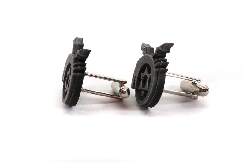 May include: A pair of black cufflinks with a silver metal clasp. Each cufflink features a raised, circular design with a star of David and wings.