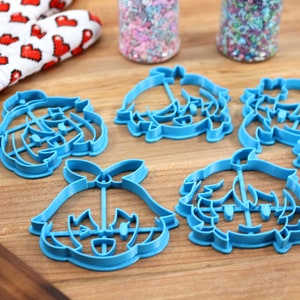 May include: Five blue plastic cookie cutters shaped like cartoon characters. The cutters are on a wooden surface.