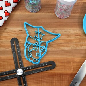 May include: A blue plastic cookie cutter in the shape of a cartoon character's face sits on a wooden surface. A blue spatula, a black measuring tool, and jars of sprinkles are also visible. A red and white heart-patterned oven mitt is in the background.