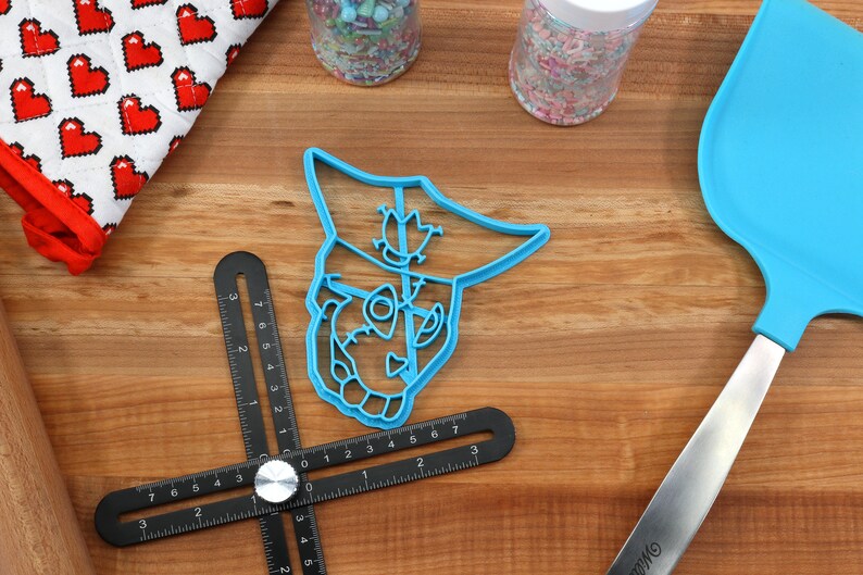 May include: A blue plastic cookie cutter in the shape of a cartoon character's face sits on a wooden surface. A blue spatula, a black measuring tool, and jars of sprinkles are also visible. A red and white heart-patterned oven mitt is in the background.