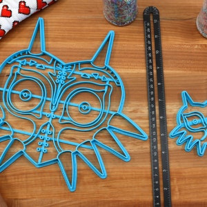 May include: Two blue plastic cookie cutters shaped like the Majora's Mask from the Legend of Zelda video game. The larger cutter is about 7 inches wide and the smaller cutter is about 4 inches wide.