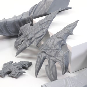 Pode incluir: Cinco pe&ccedil;as impressas em 3D de cor cinza com designs intrincados, que se assemelham a asas ou armaduras. As pe&ccedil;as est&atilde;o sobre uma superf&iacute;cie branca.
