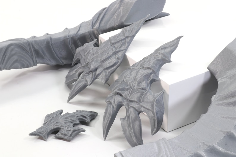 Pode incluir: Cinco pe&ccedil;as impressas em 3D de cor cinza com designs intrincados, que se assemelham a asas ou armaduras. As pe&ccedil;as est&atilde;o sobre uma superf&iacute;cie branca.