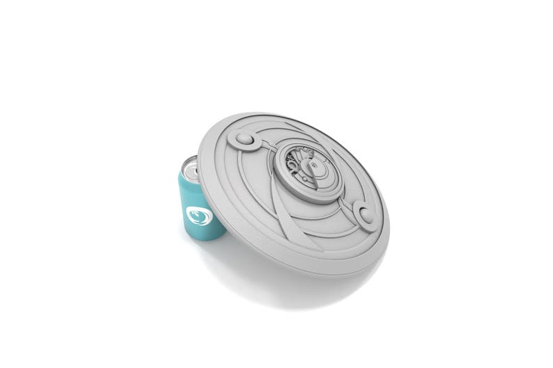 May include: A light gray, circular shield-shaped object with intricate, raised designs, partially covering a teal can with a white logo. The shield has a central, detailed emblem and curved accents. The can is cylindrical.