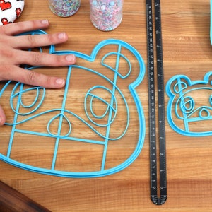 May include: Two blue plastic cookie cutters shaped like the Animal Crossing character, Isabelle. The larger cutter is about 7 inches wide and the smaller cutter is about 4 inches wide. The cutters are on a wooden surface.