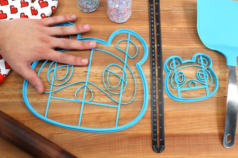 May include: Two blue plastic cookie cutters shaped like the Animal Crossing character, Isabelle. The larger cutter is about 7 inches wide and the smaller cutter is about 4 inches wide. The cutters are on a wooden surface.
