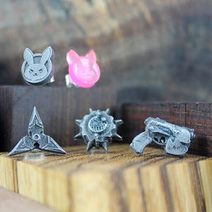 May include: Five small, silver-colored, 3D printed charms. The charms are shaped like a bunny, a star, a gun, a spiky ball, and a heart. The heart charm is pink.
