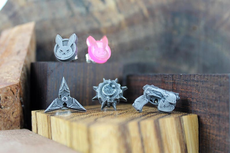May include: Five small, silver-colored, 3D printed charms. The charms are shaped like a bunny, a star, a gun, a spiky ball, and a heart. The heart charm is pink.